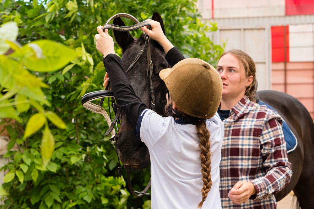 Woman and young rider collaboratively adjusting horse's bridle.