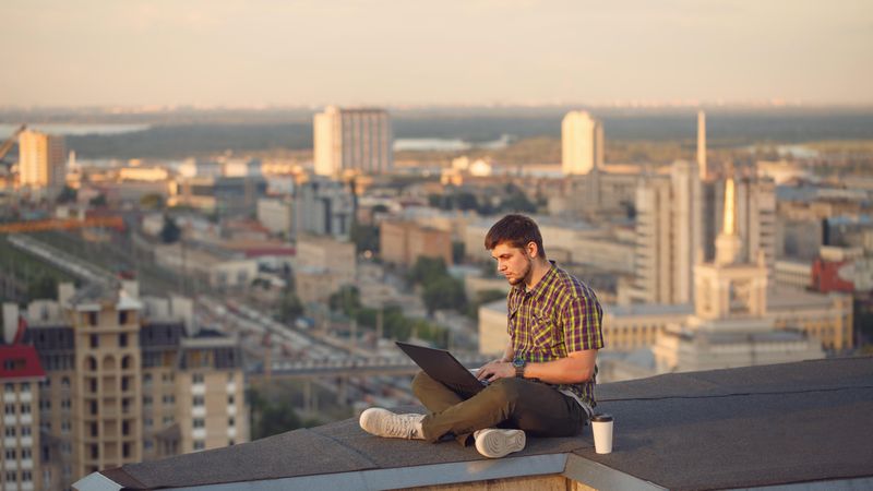 Man sitting crossed legged on roof over looking city working on laptop with takeaway drink