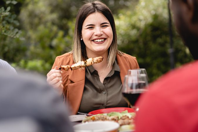 Woman smiles and chats with multi-ethnic group of friends eating meat skewers in the backyard