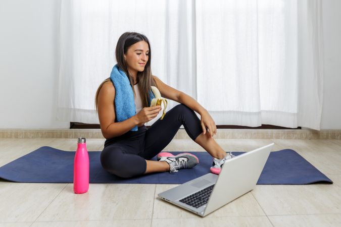Young woman eating a banana and sitting beside laptop on yoga mat