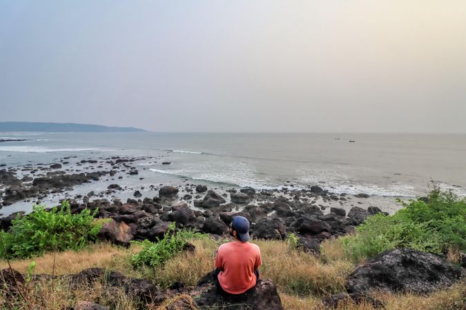 Back view of man in red shirt sitting on rock near seashore