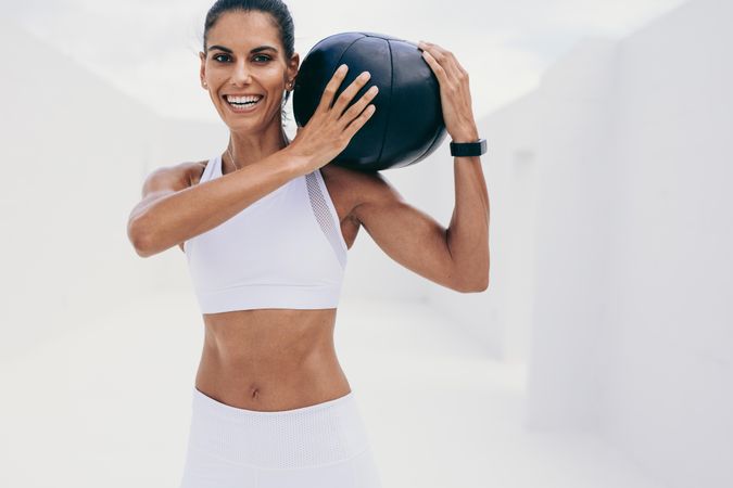 Smiling woman in fitness wear doing workout with a medicine ball on her shoulder