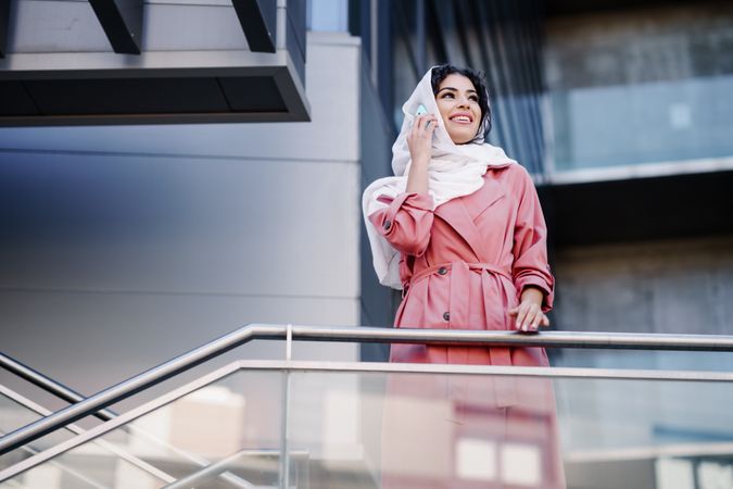 Female in headscarf and pink trench coat standing on staircase speaking on phone, copy space
