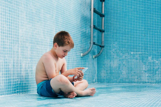 Boy with smartphone sitting on bottom of empty pool