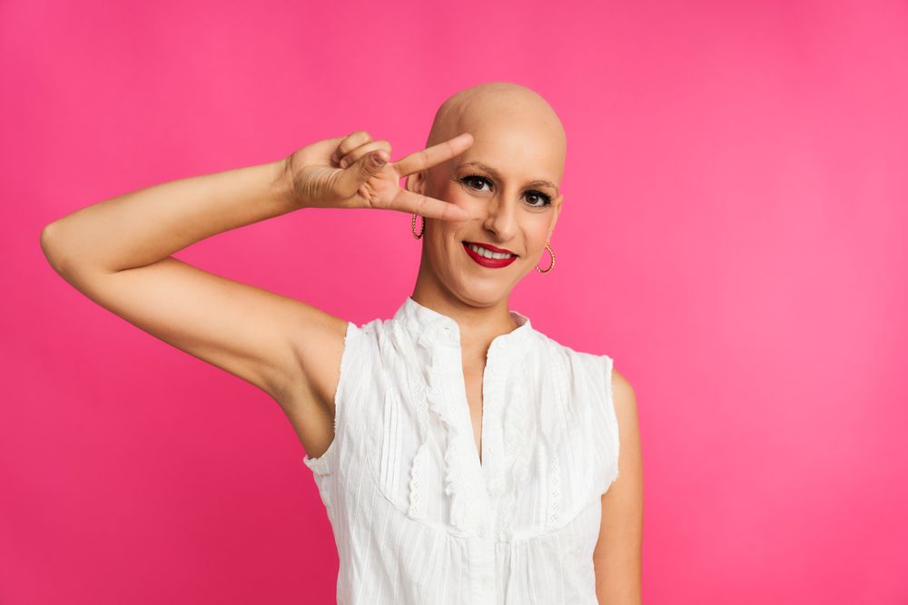 a woman suffering from cancer with a shaved head and a victory gesture on a pink background