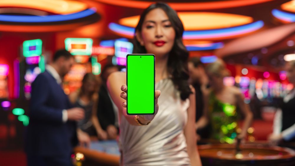 Modern Casino Setting: Gorgeous Asian Brunette Woman Posing with a Mobile Phone with a Green Screen Placeholder Space for Advertising
