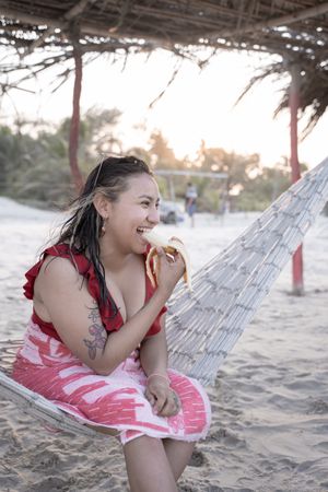 A young hispanic woman is eating a banana on a hammock during sunset