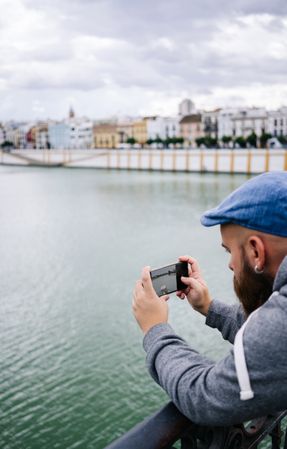 Guy on vacation in casual clothing making photo of bay on smartphone at quay