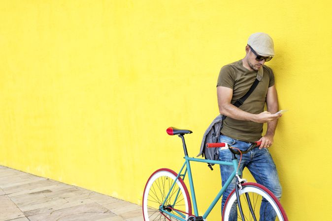 Male in hat and sunglasses standing next to yellow wall with bike and texting on phone, copy space