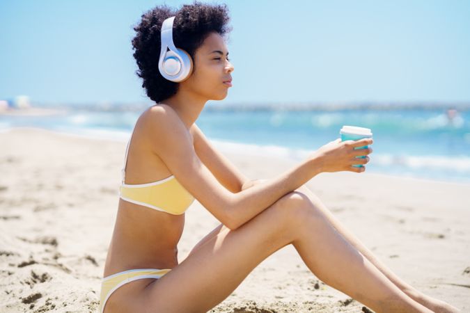 Woman in yellow bikini sitting on beach with coffee cup