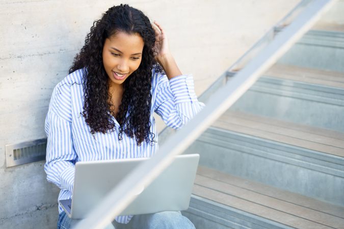 A young woman is focused on her laptop while sitting on the stairs, showing inspiration