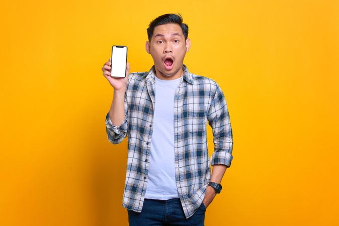 Surprised Asian male with hand in pocket and showing blank screen of smart phone