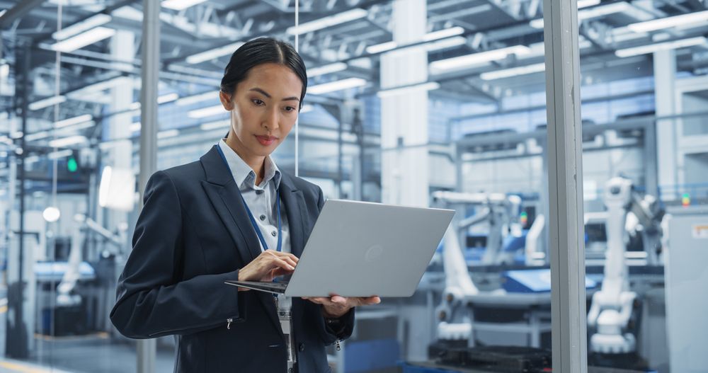 Thoughtful Asian Engineer Monitoring and Analyzing Conditions at a Pristine Modern Electronics Factory with Automated Robot Hands Working with the Help of Artificial Intelligence Software
