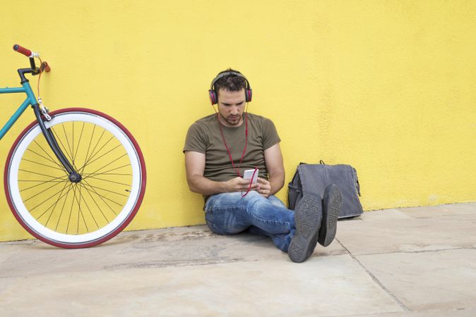 Male sitting in front of yellow wall next to bike listening to music on smartphone