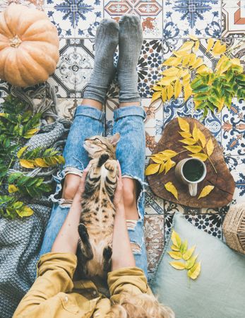 Woman with cat on lap sitting on colorfully tiled balcony with fall leaves, squash, and mug