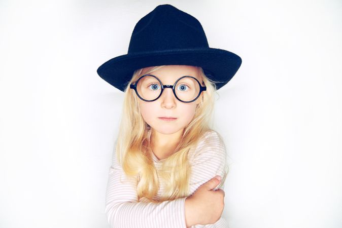 Serious blonde girl with pursed lips wearing hat and glasses with her arms crossed