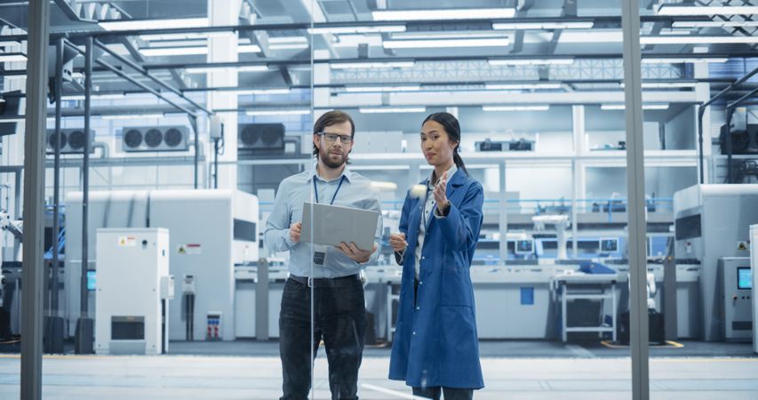 Man and Asian Female Having a Conversation at an Industrial Facility. Advanced Factory Employees Working Together, Using Laptop Computer for Supervising Work Process in a Facility