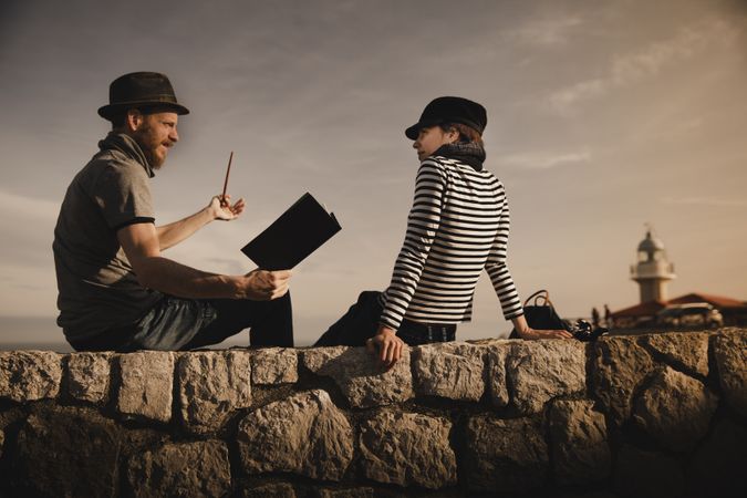Man with pipe near woman with book on stones near lighthouse