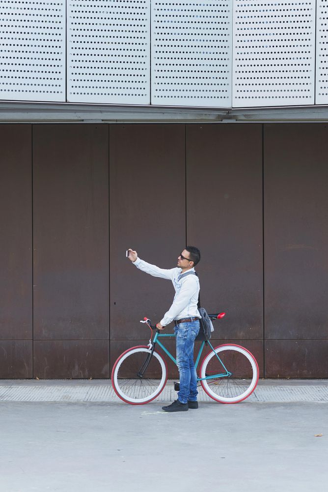 Male in sunglasses standing with red and green bicycle taking selfie parked against a brown wall