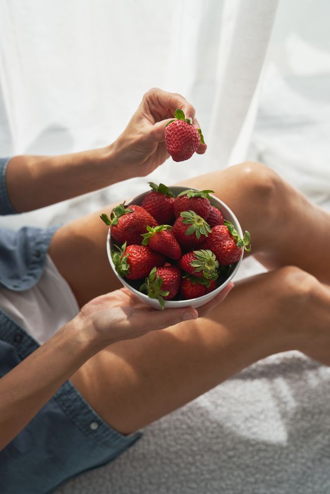 Unrecognizable female taking strawberry from bowl while sitting