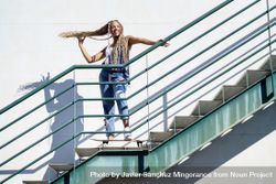 Female in denim overalls standing on skateboard on stairs playing with her hair 0KwPD0
