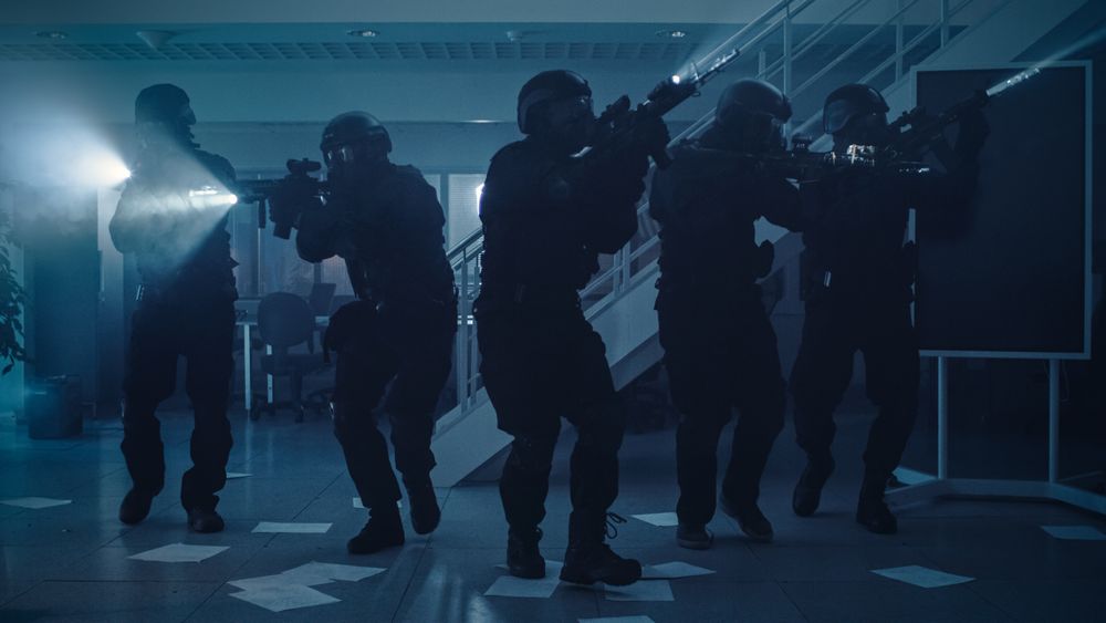 Masked Team of Armed SWAT Police Officers Move in a Hall of a Dark Seized Office Building with Desks and Computers.