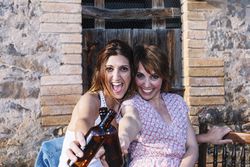 Two cheerful women holding bottles of beer and smiling while sitting outdoors