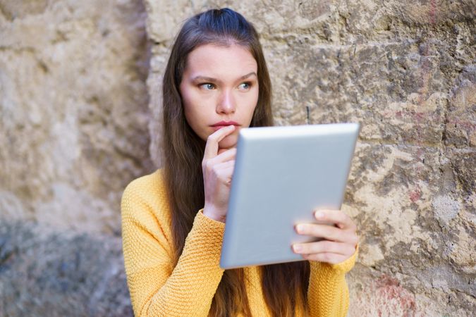 A young woman is deeply engrossed in the tablet screen while sitting against a stone wall