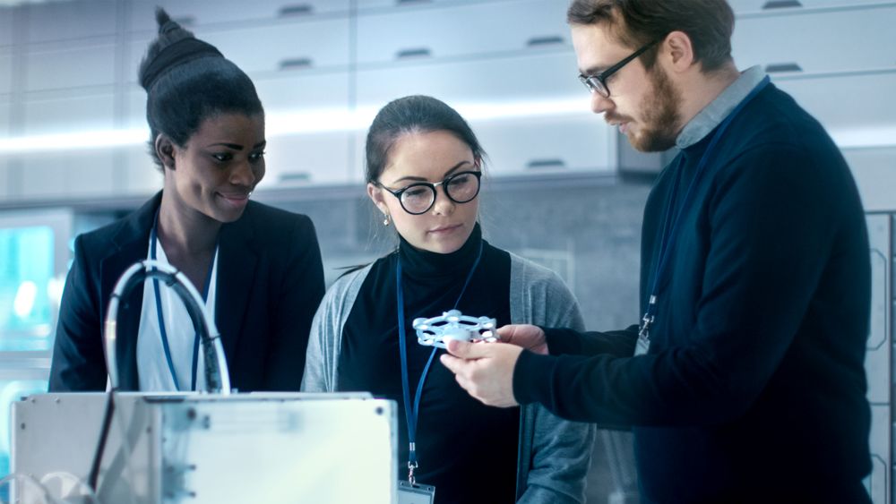 Three Very Bright and Young Multiethnic Students Discussing 3D Printed Mechanism in a New Modern Laboratory.
