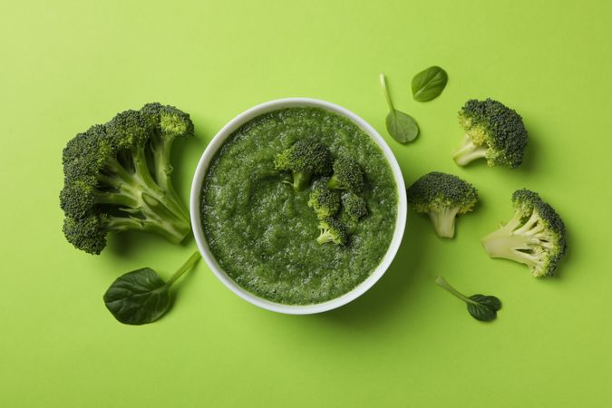 Top view of bowl of broccoli soup surrounded with vegetables on green table