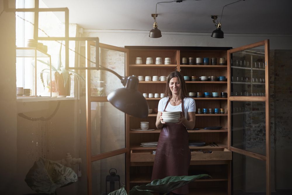 Smiling woman in leather apron holding handmade plates in her shop