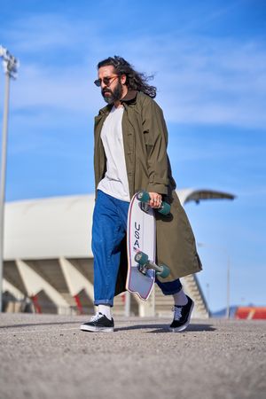 Stylish adult man in raincoat walking on street with skateboard