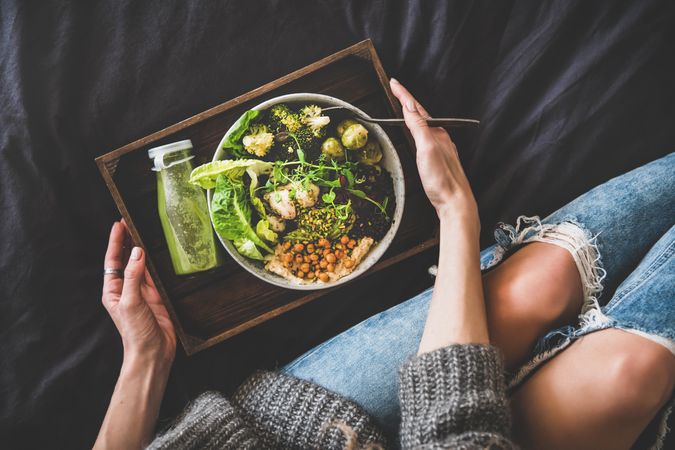 Woman in jeans sitting with wooden tray of vegetarian bowl with smoothie on side