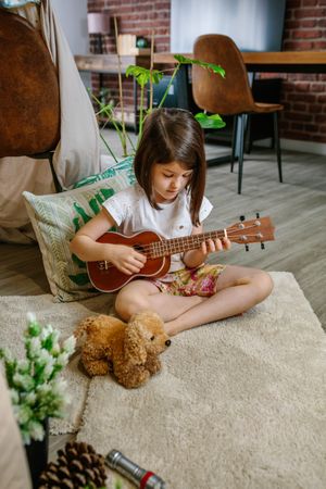 Little girl playing ukulele sitting on carpet next to handmade shelter tent in living room at home