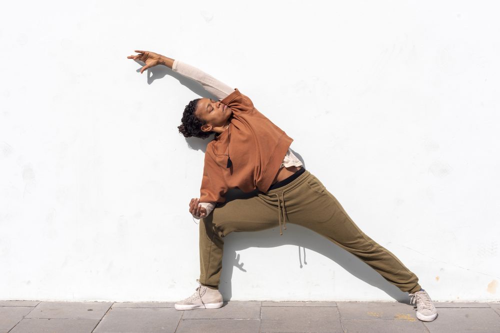 Stylish young female runner in sporty clothes stretching before running against wall