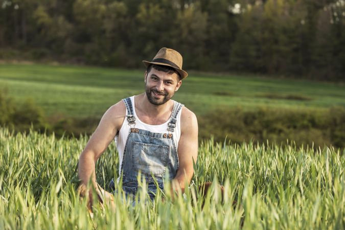 Bearded smiling young man wearing a hat sitting in field while looking camera