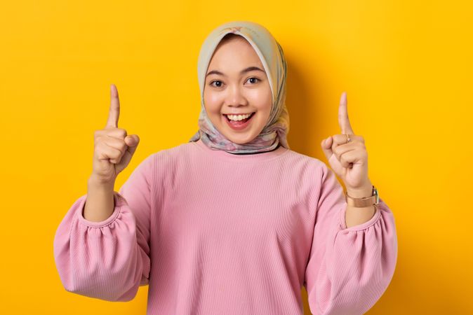 Smiling Muslim woman on the phone and pointing two fingers upwards holding smart phone