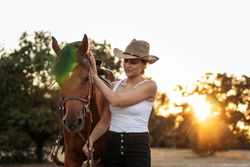Young woman in hat walking with her horse in the countryside at sunset