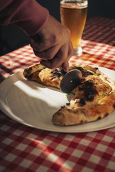 Calzone being sliced with pizza cutter on plate at checkered table with beer