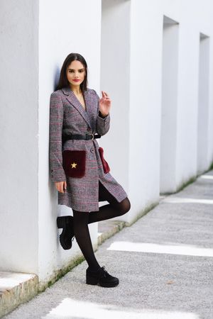 Female leaning on bright wall outside in belted winter coat dress