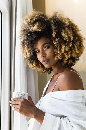 Dreamy Black woman with cup of drink near window