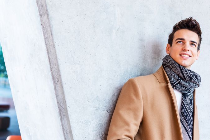 Smiling man in camel coat and scarf looking away leaning on wall outside