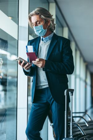 Man with face mask standing at airport waiting area using his mobile phone