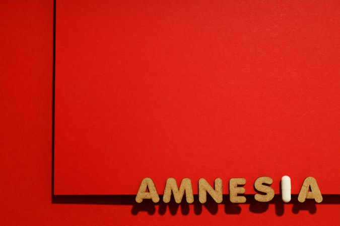 Cork letters of the word “Amnesia” with pill along bottom of red background with copy space
