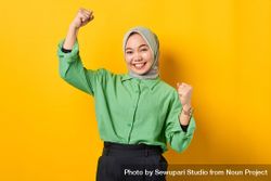 Smiling Muslim woman in headscarf and green blouse making two fists in victory 5azNP4
