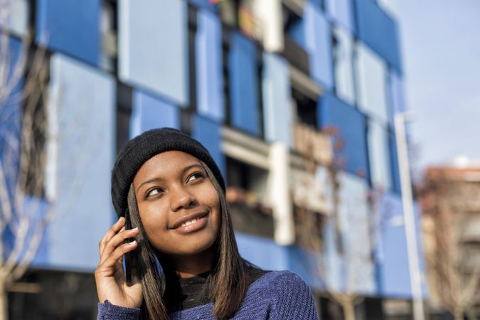 Female in wool hat and blue sweater chatting on cell phone and looking to her side