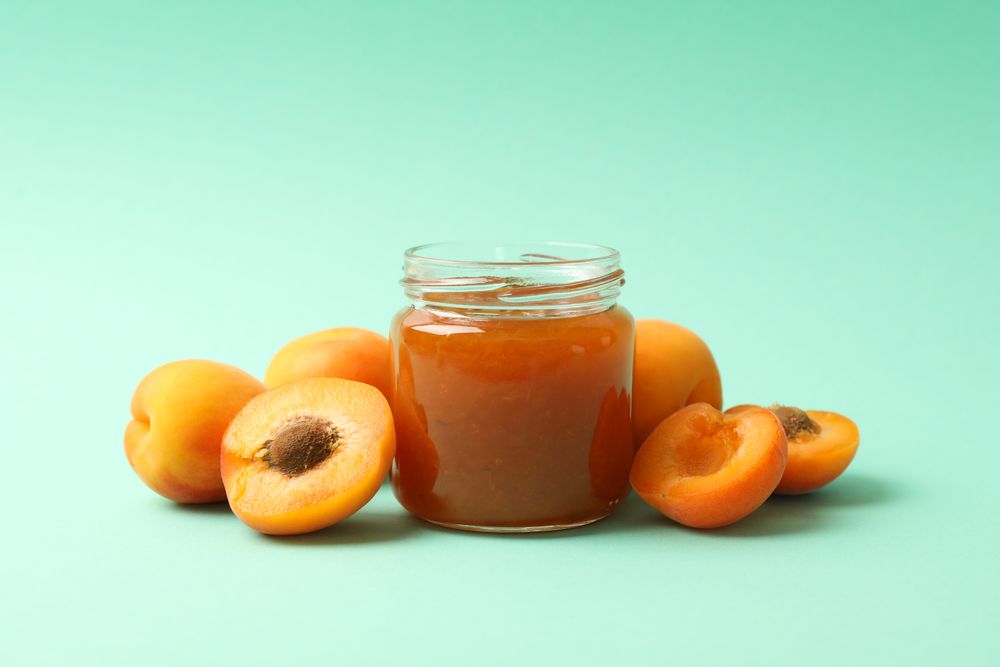 Glass jars with apricot jam and ingredients on mint background, space for text