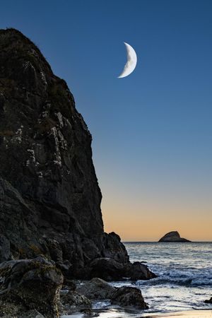 Crescent moon above quiet rocky beach at dusk, vertical composition