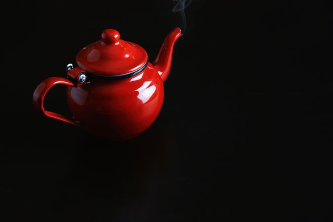 Enamel red tea pot on dark table with steam rising from spout