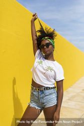 Young Black woman with sunglasses standing against yellow wall and playing with hair 5Q28rm